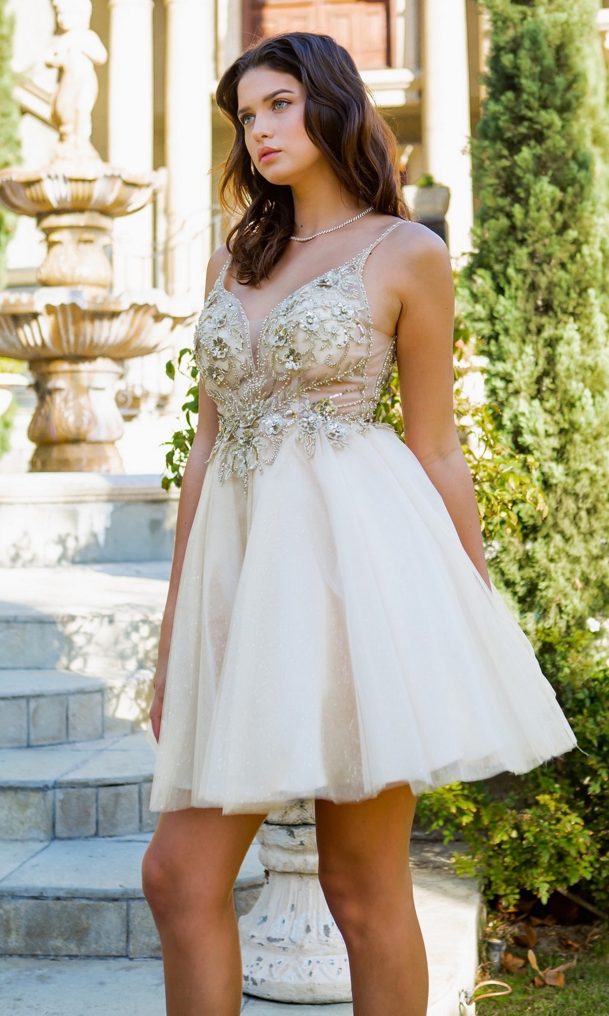 A woman stands outdoors near steps and a fountain wearing the Short Ball-Gown-Style Homecoming Dress 5112J, a sleeveless, light-colored dress with floral embellishments.